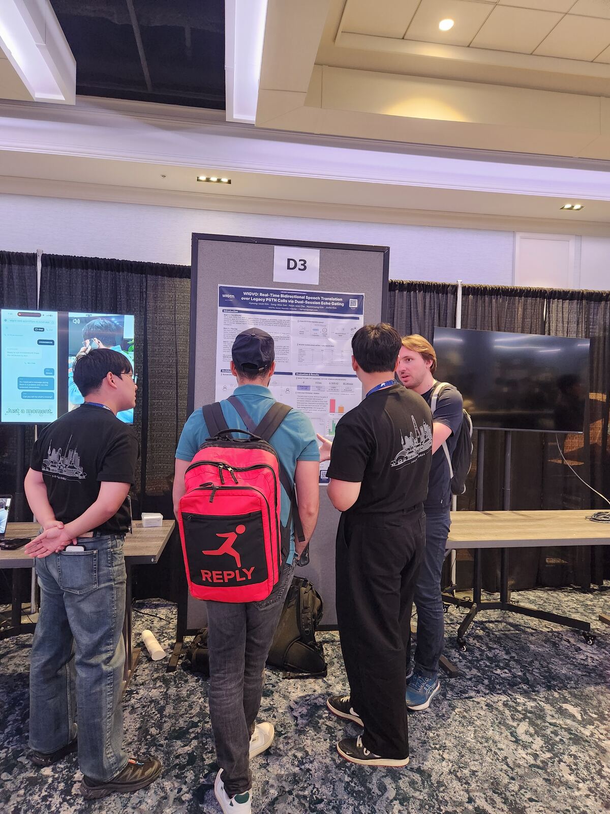 Wide view of the WIGTN booth showing the poster, the monitors and the demo table.