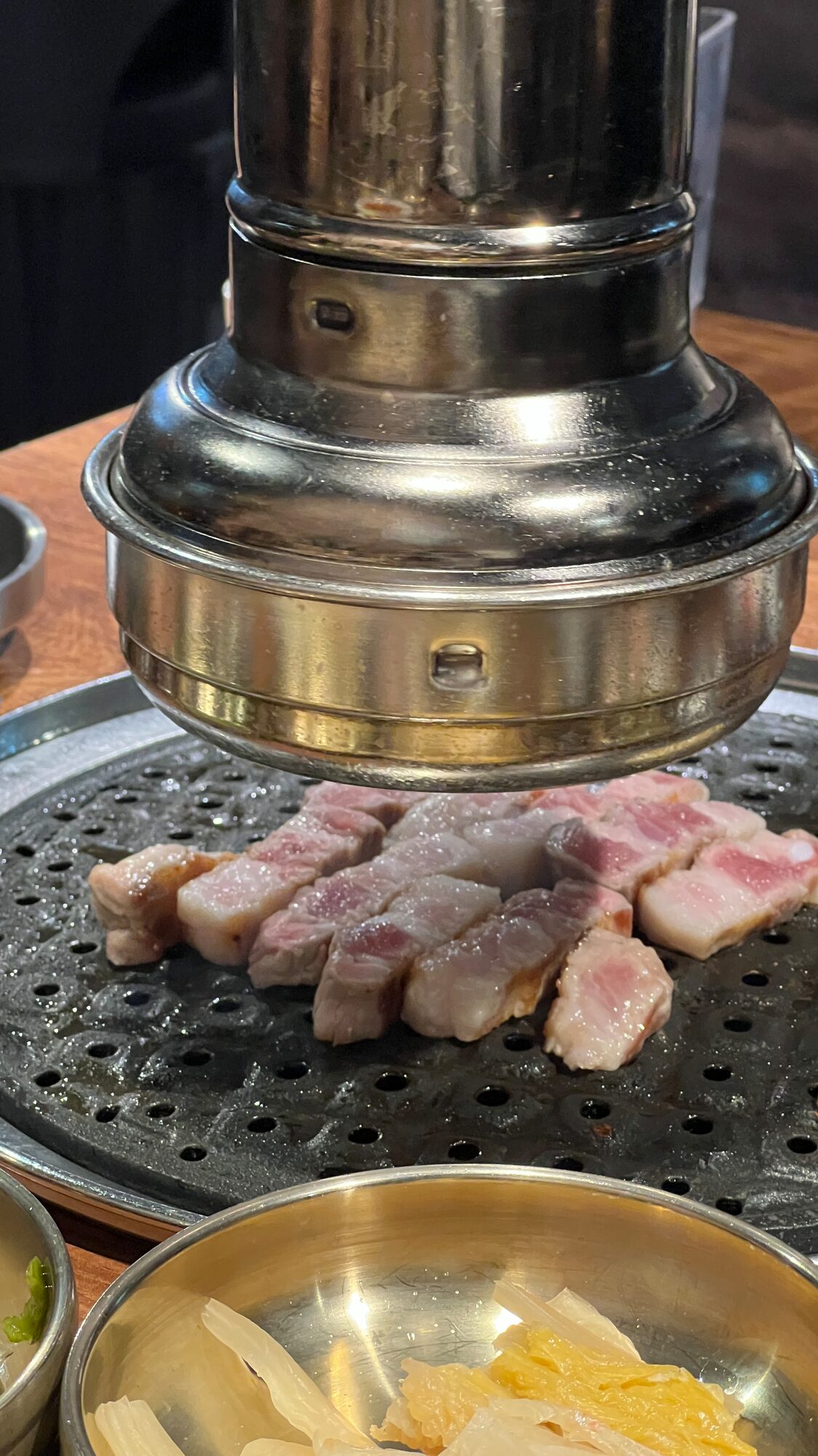 Samgyeopsal cooking on a perforated grill plate under a steel flue, with a bowl of pickled radish in the foreground.