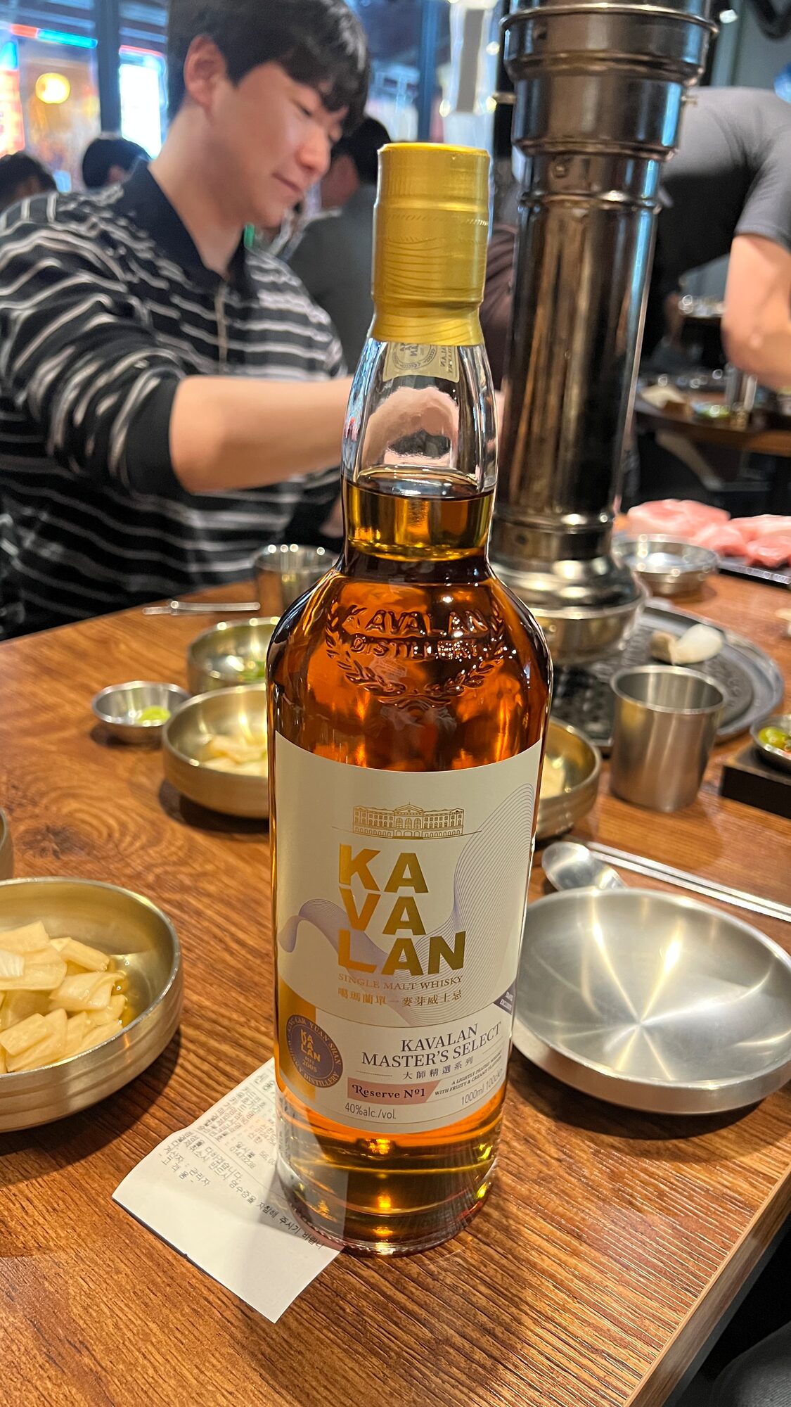 A bottle of Kavalan Master's Select on a wooden restaurant table beside brass banchan bowls and a grill flue, with a teammate seated behind it.