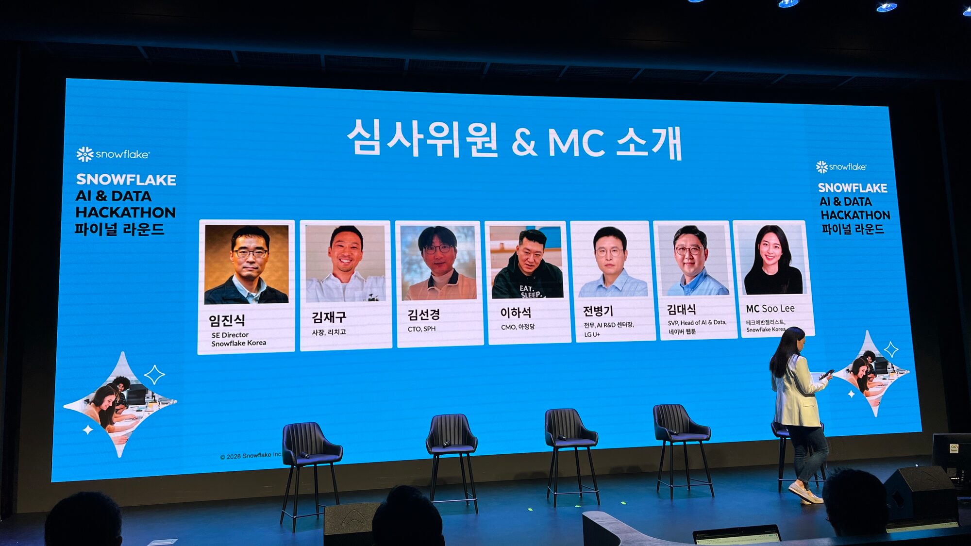 The "심사위원 & MC 소개" slide on the hackathon stage, showing seven headshots in a row with names and titles: Snowflake Korea's SE Director, RichGo's CEO, SPH's CTO, AJD's CMO, LG U+'s AI R&D centre head, Naver Webtoon's SVP of AI and Data, and the MC.