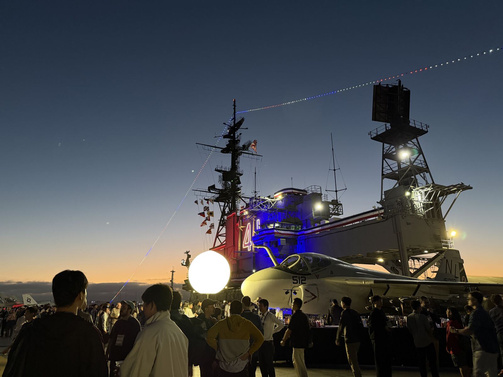 The conference reception on the USS Midway flight deck at sunset, with attendees gathered around the bar beside a parked aircraft.