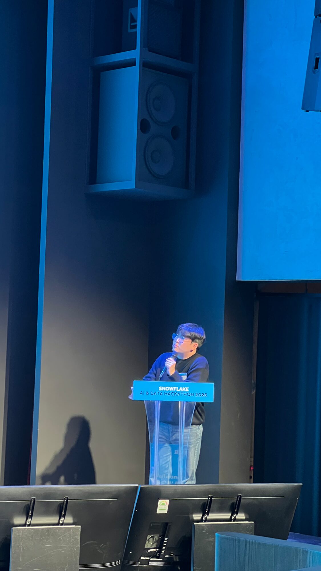 A WIGTN presenter alone at the acrylic podium on the Snowflake hackathon stage, lit blue, speaking into a handheld microphone.