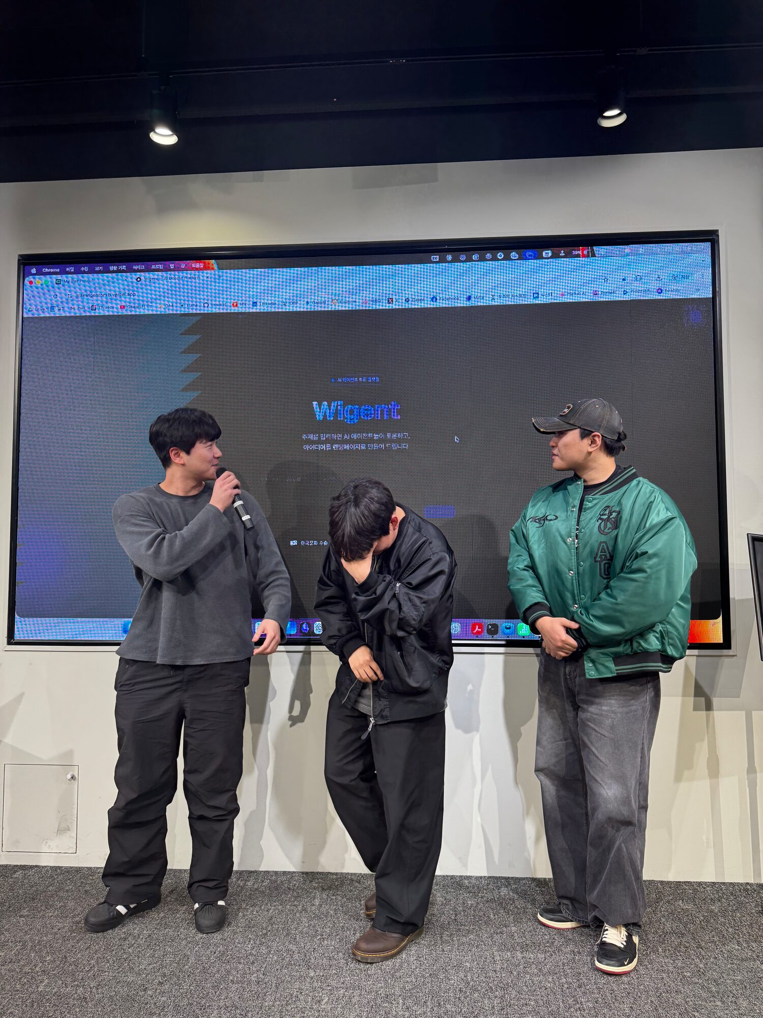 Three WIGTN engineers presenting WIGENT on stage at Build with TRAE Seoul, with the Wigent topic-entry screen open in a browser on the wall display behind them.