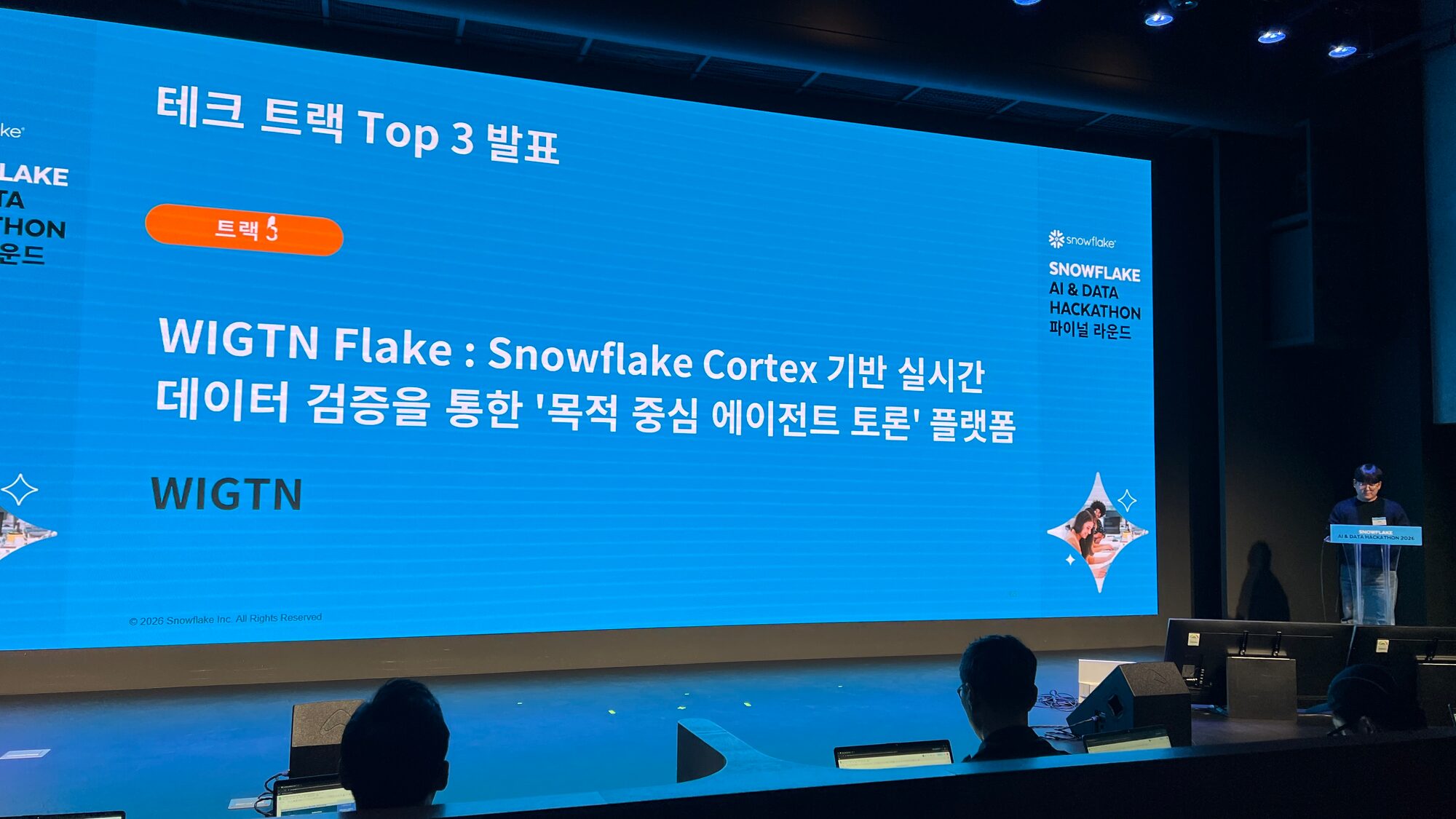 The Tech Track Top 3 announcement slide on stage, reading "WIGTN Flake : Snowflake Cortex 기반 실시간 데이터 검증을 통한 '목적 중심 에이전트 토론' 플랫폼" under a "트랙 3" badge.